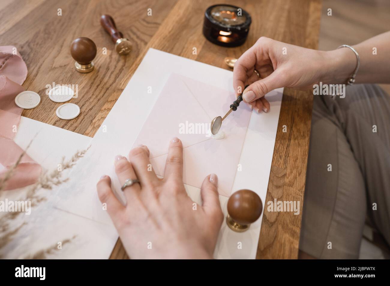 Gepflegte Hände einer nicht erkennbaren Frau, die flüssiges weißes Wachssiegel mit Löffel auf den pinken Grußumschlag auf den Schreibtisch gießt. Stockfoto