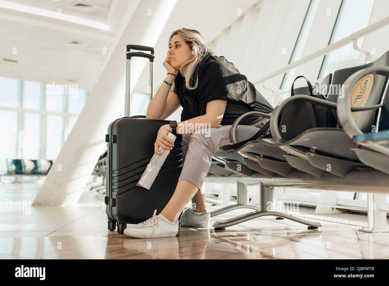 Müde sitzende Frau, die den Kopf an die Hand legt, sich in der Lounge auf den Koffer lehnt und ein zusammengerolltes Magazin hält. Flugverspätung. Stockfoto