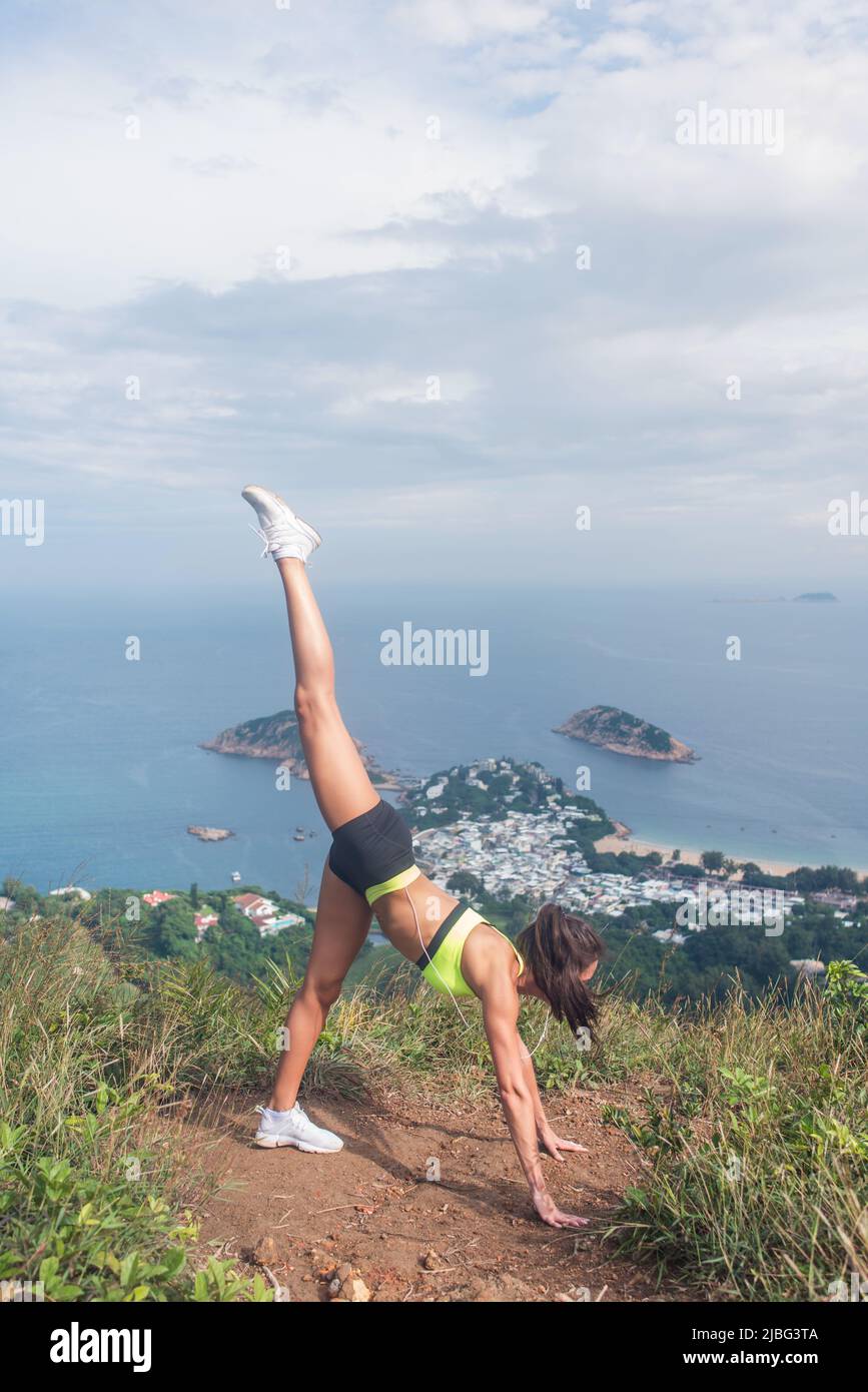 Junge Frau, die im Stehen geteilte Yoga-Übungen macht, steht auf dem Berggipfel gegen bewölkten Himmel, Meer und Inseln im Hintergrund. Fit Mädchen ausstrecken outdoo Stockfoto