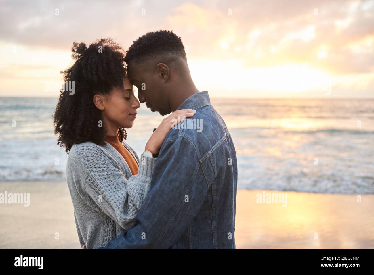 Junges multiethnisches Paar, das bei Sonnenuntergang am Strand in den Armen steht Stockfoto