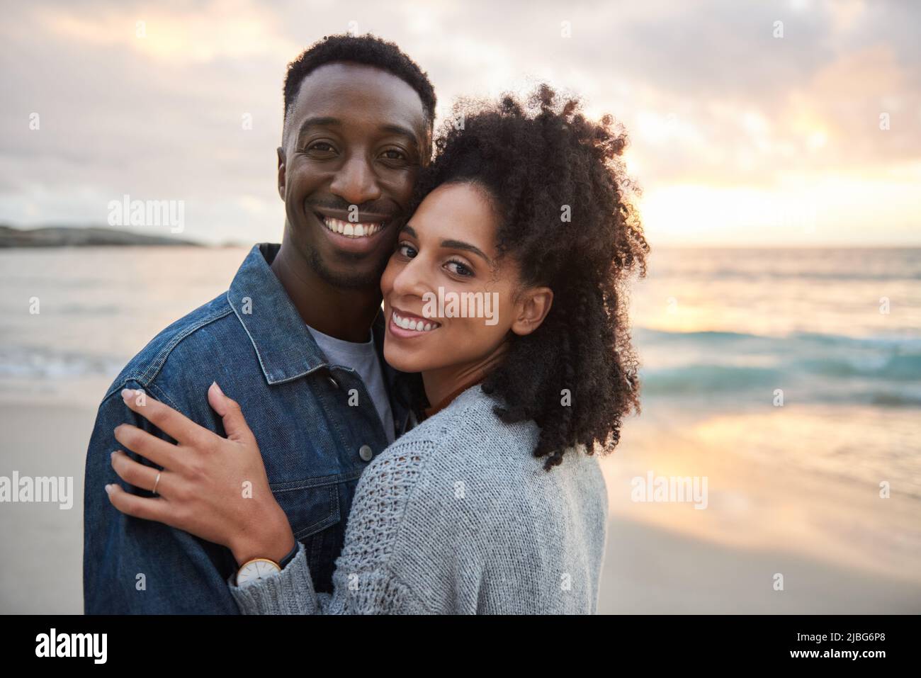 Lächelndes multiethnisches Paar, das Arm in Arm am Strand bei Sonnenuntergang steht Stockfoto