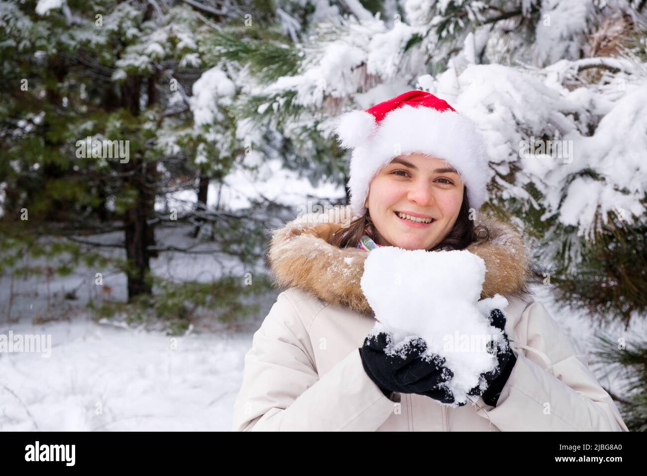 Eine Frau im Weihnachtsmann-Hut hält einen Schneeball in ihren Händen in einem schneebedeckten Winterwald, ein Ort für Text Stockfoto