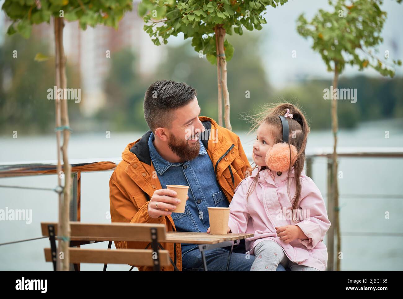 Mann hält eine Tasse Kaffee und lächelt, während er Zeit mit dem Kind im Café im Freien verbringt. Glücklicher Vater und Tochter sitzen am Tisch auf der Straße im neuen Stadtbezirk. Konzentrieren Sie sich auf Mann und kleines Mädchen. Stockfoto