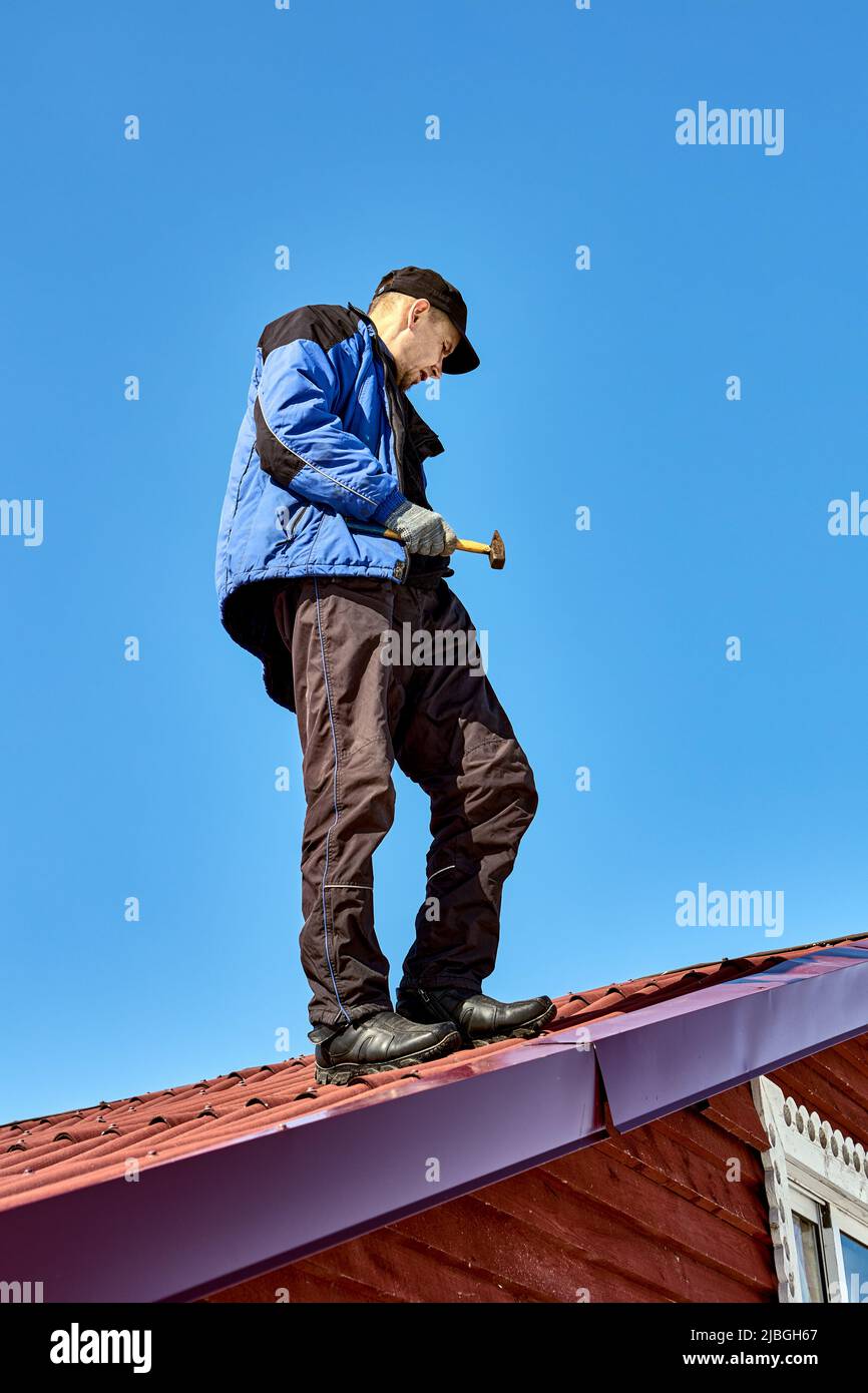 Der Dachdecker steht mit Hammer in der Hand vor dem Hintergrund des blauen Himmels auf dem Dach. Stockfoto