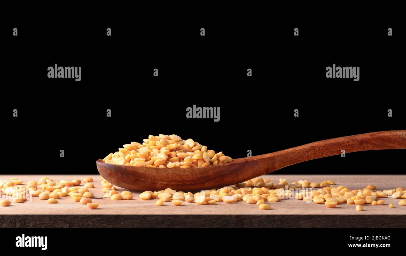 Stapel gelber Erbsen in einem Holzlöffel und verstreute Erbsen auf der Tischplatte, geschälte und gespaltene Erbsen, isoliert auf schwarzem Hintergrund, Platz kopieren Stockfoto