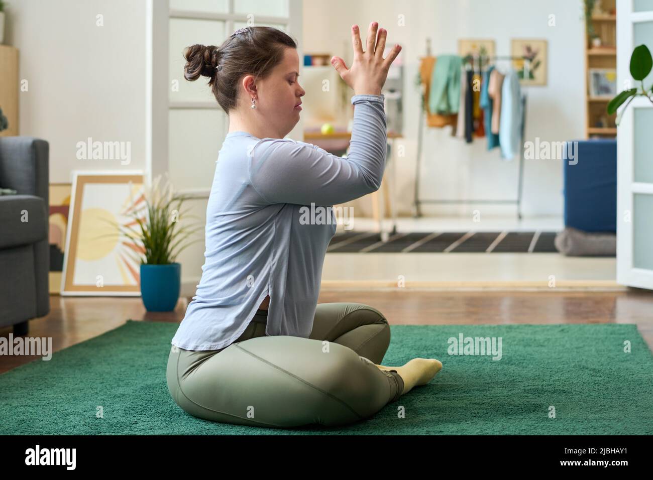 Junge Frau in aktivem Ohr, die die Hände vor ihrem Gesicht zusammengehalten hat, während sie in Lotusposition sitzt und zu Hause Yoga praktiziert Stockfoto