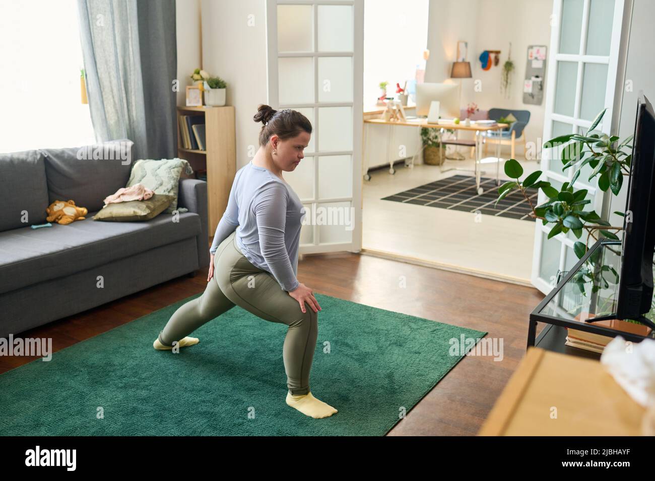 Aktives Mädchen mit geistiger Behinderung macht Dehnungsübungen auf dem Boden des Wohnzimmers vor dem fernsehbildschirm, während es Yoga praktiziert Stockfoto