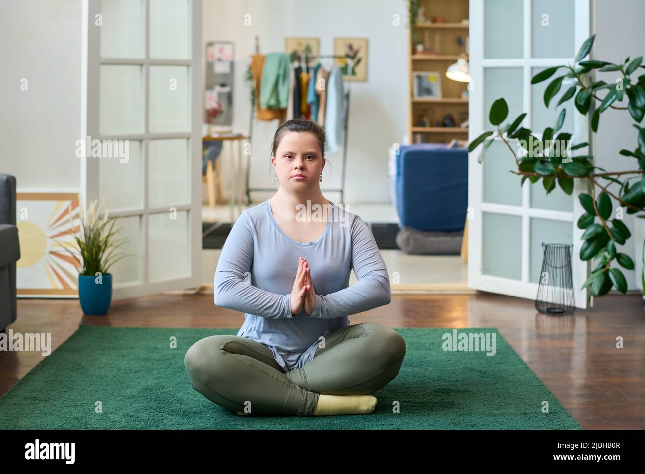 Junge Frau mit Behinderung sitzt in Lotuspose auf dem Boden des geräumigen Wohnzimmers und schaut während des Yoga auf die Kamera Stockfoto