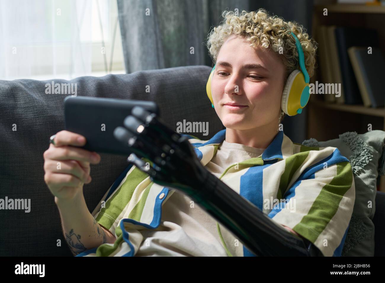 Glückliches Mädchen mit myoelektrischem Arm, das Online-Film auf dem Smartphone sieht, während es sich auf dem Sofa im Wohnzimmer entspannt Stockfoto