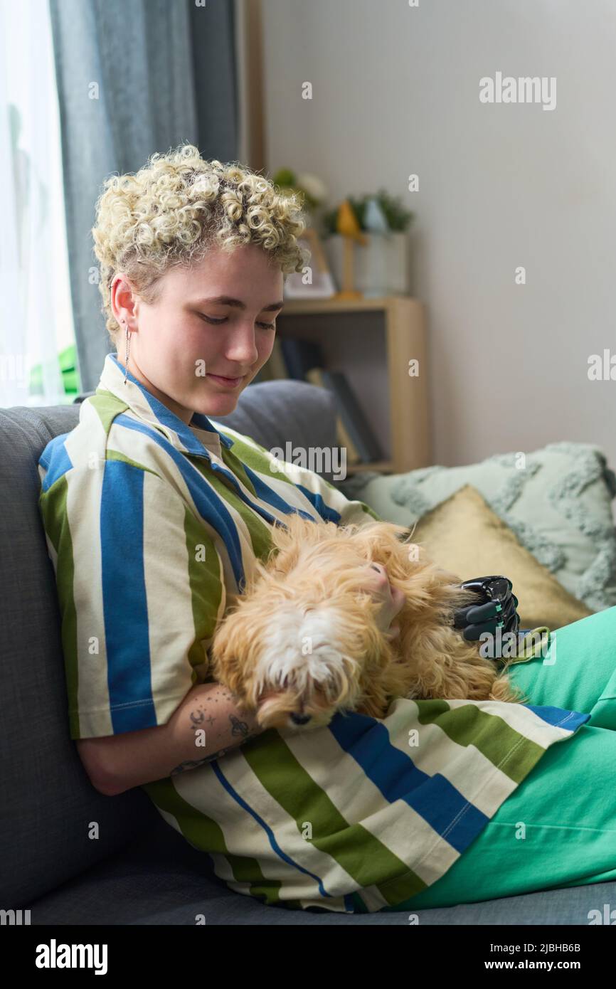 Glückliches Mädchen mit körperlicher Behinderung spielt mit niedlichen reinrassigen Hund, während Sie sich auf der bequemen Couch im Wohnzimmer in Ruhe entspannen Stockfoto