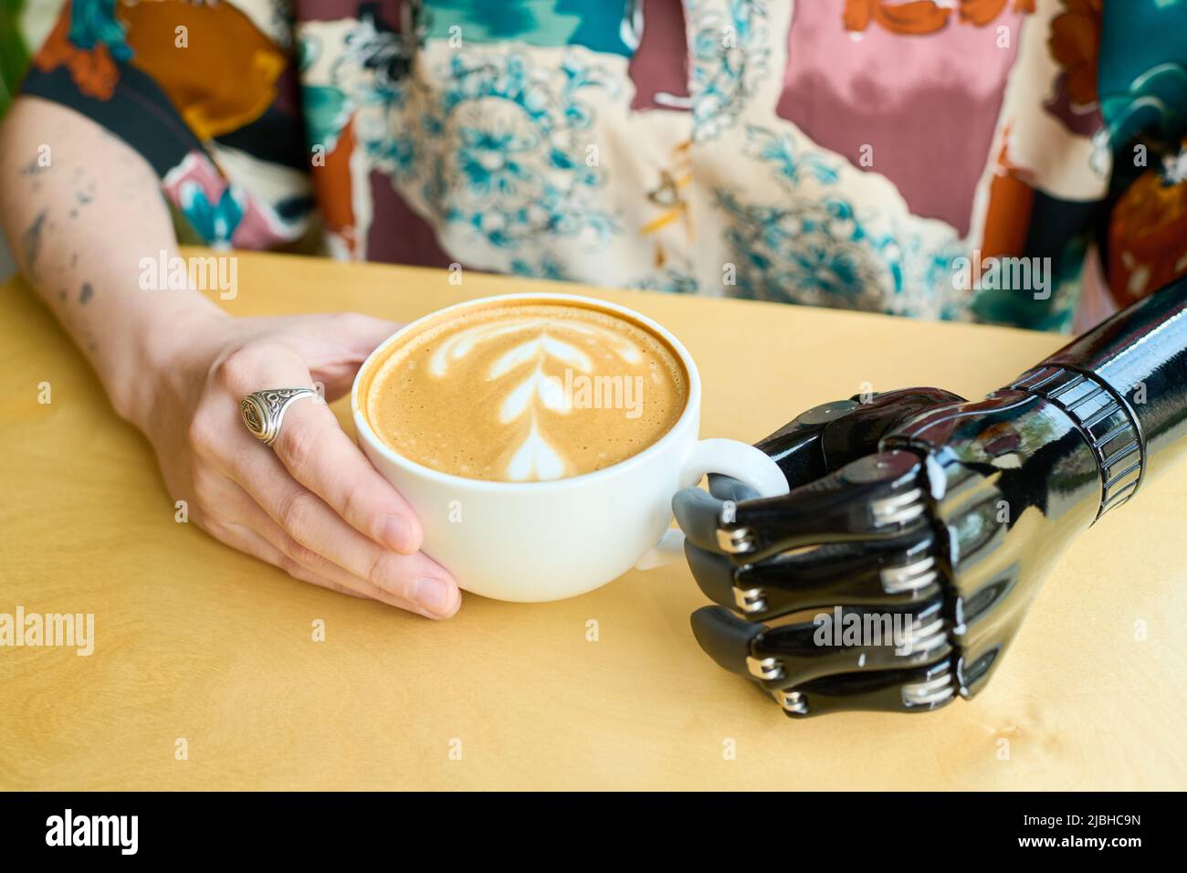 Junge, moderne Frau mit myoelektrischem Arm, die im Café eine Tasse Cappuccino hält, der mit flauschigem Milchschaum dekoriert ist Stockfoto