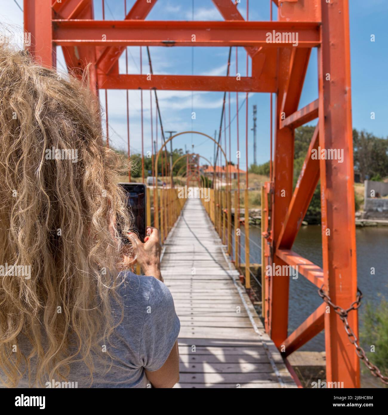 Rückansicht einer blonden Frau, die mit dem Smartphone eine Fußgängerhängebrücke fotografiert hat. Stockfoto