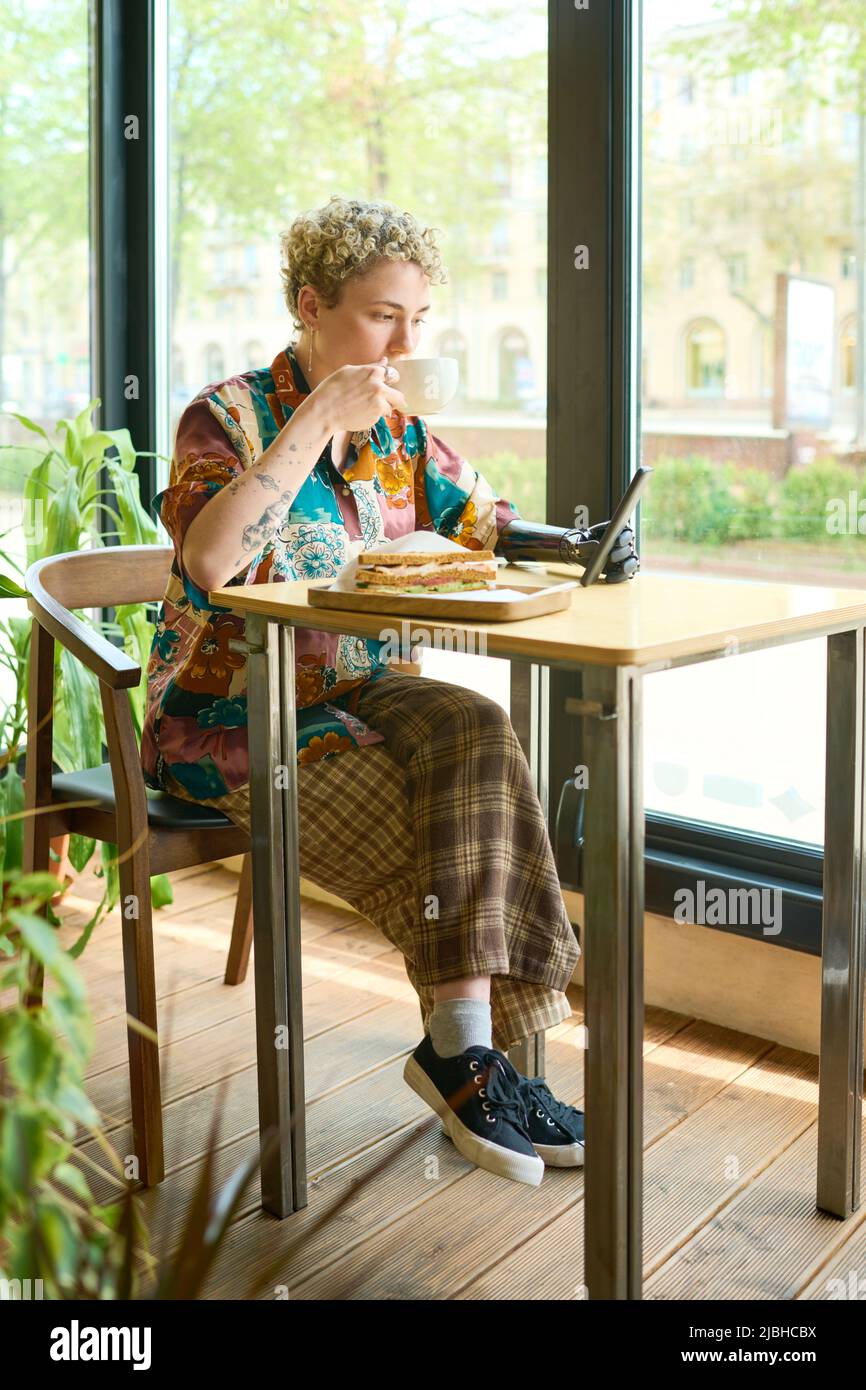 Junge, erholsame Frau mit myoelektrischem Arm, die Smartphone benutzt und Kaffee mit Sandwich zum Frühstück getrunken hat, während sie am Tisch im Café sitzt Stockfoto
