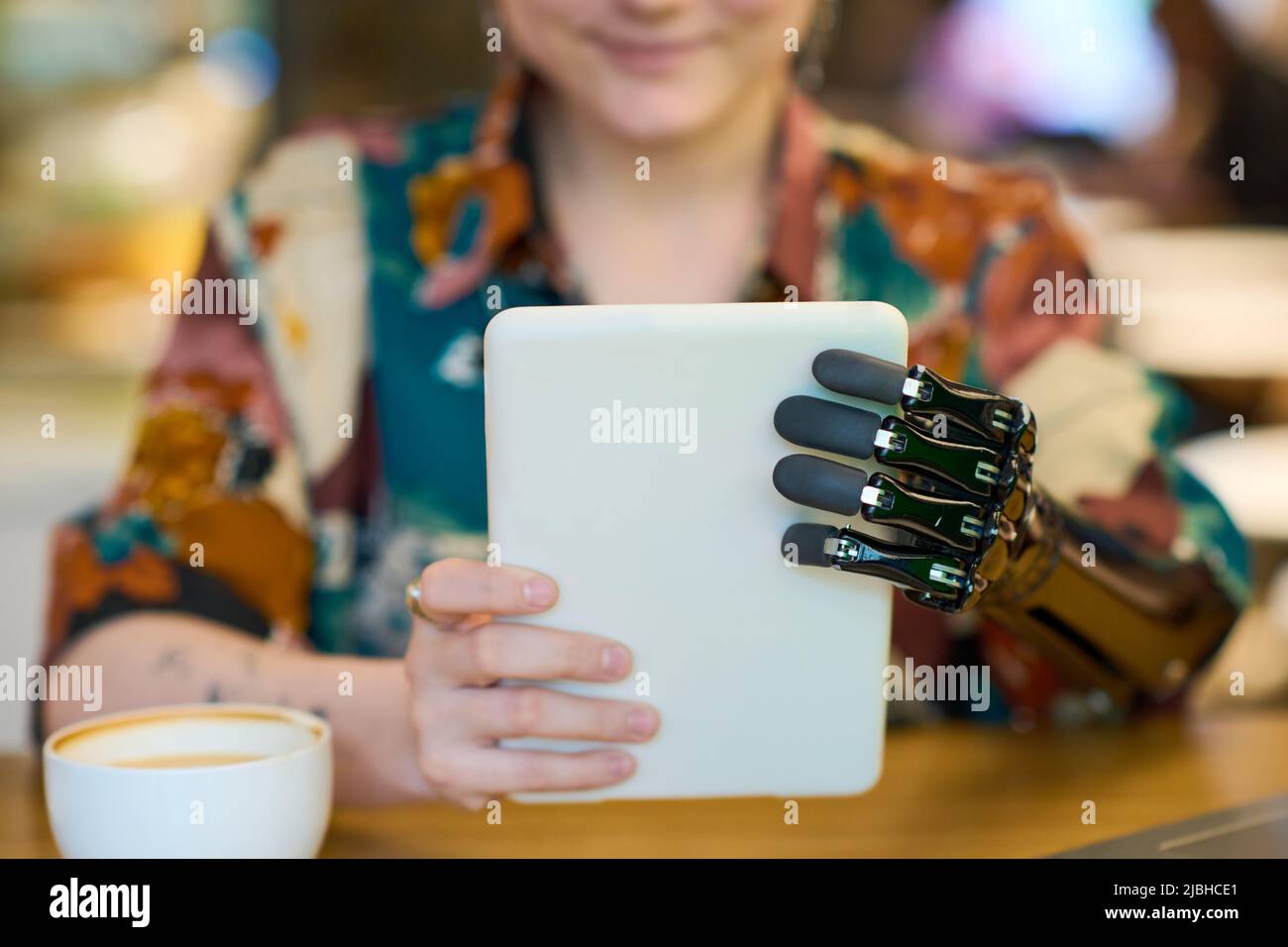Hände einer jungen Frau mit myoelektrischem Arm, die die Tablette vor sich hält, während sie im Café am Tisch sitzt und Kaffee getrunken hat Stockfoto