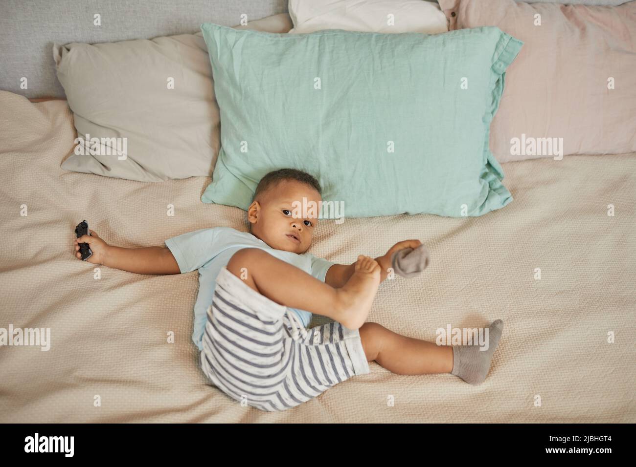 Aus dem hohen Winkel sieht man das niedliche schwarze Kleinkind, das in pastellfarbenen Laken auf dem Bett liegt und mit Socken spielt und die Kamera anschaut Stockfoto