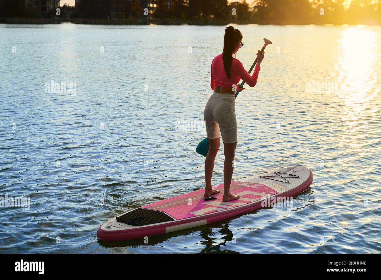 Sportliche junge Frau mit langem Paddel in den Händen, die am Abend auf dem Brett schwebt. Gesund und fit Brünette Training auf Stadtsee mit erstaunlichen Sonnenuntergang auf dem Hintergrund. Stockfoto