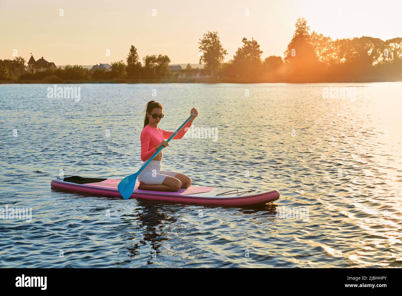 Starke und gesunde Frau in Sportbekleidung und Sonnenbrille schwebt auf dem Brett mit Sommeruntergang auf dem Hintergrund. Junge Brünette genießen Wassersport während der Abendzeit. Stockfoto