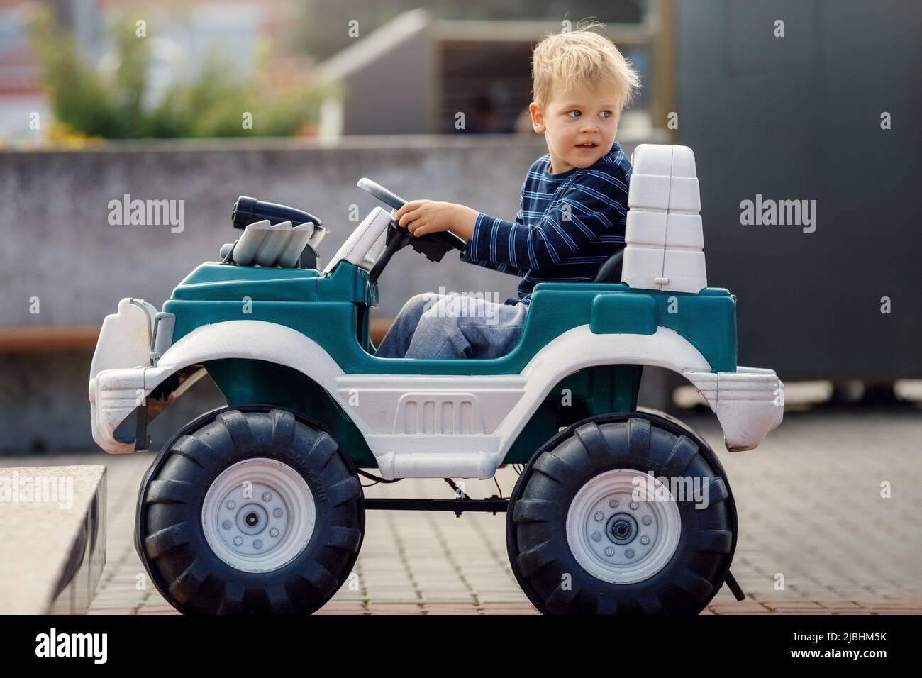 Lustige junge Autofahrer mit dem Lenkrad. Dreijähriger Junge in einem blauen Pullover in einem grünen Spielzeugauto in der Stadt. Kleiner Junge fährt große Spielzeugauto und Stockfoto