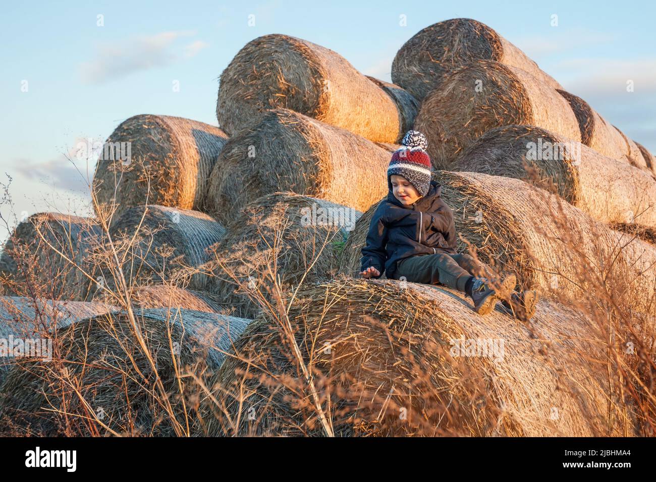 Ein Kind, das warme Herbstkleidung und einen Hut trägt, sitzt im Abendlicht und spielt im Dorf auf einem Haufen goldener Strohrollen. Stockfoto