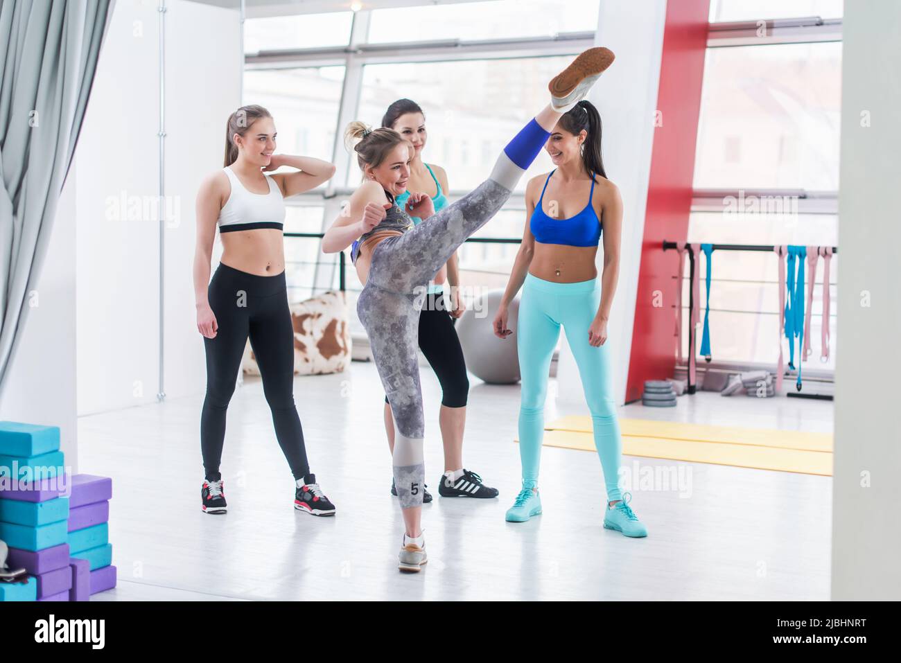 Attraktive junge Mädchen zeigt hohen Kick zu ihren Freunden, die um sie herum in Sport-BH und Leggings lächelnd entspannend nach dem Training in Sport cl Stockfoto