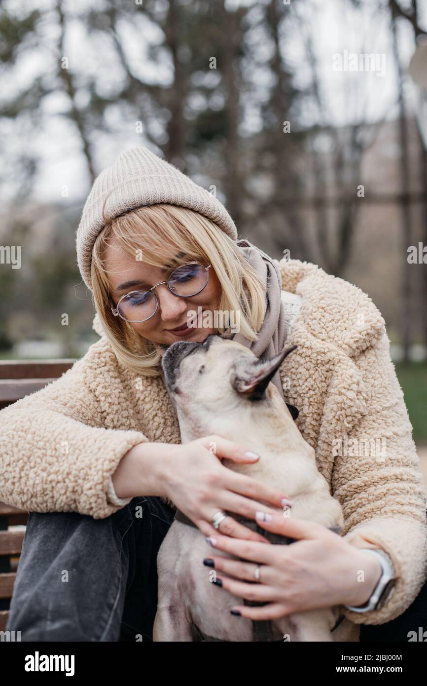 Hübsche Blondine umarmt ihren niedlichen Mops. Schöne junge Frau, die draußen mit ihrem Haustier in den Händen sitzt. Stockfoto