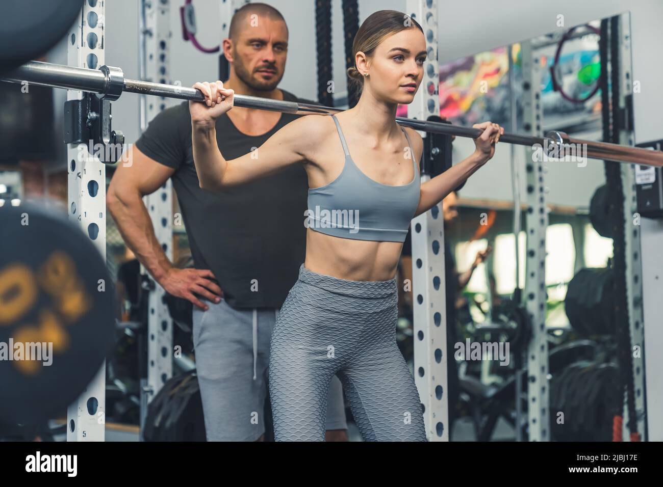 Stolze selbstbewusste kaukasische junge Erwachsene fit Frau im Fitnessstudio Gewichtheben unterstützt von ihrem persönlichen Trainer. Sportkonzept. Hochwertige Fotos Stockfoto