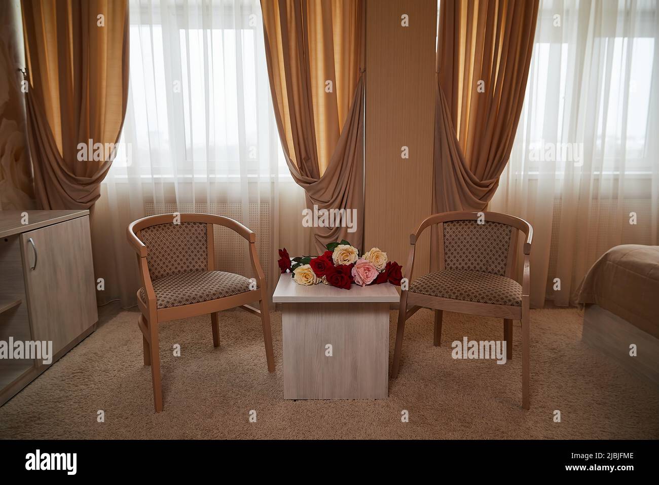 Ein Wohnzimmer mit einem hellen Teppich, zwei Sesseln, einem kleinen Tisch und einem Fenster mit verdunkelten Vorhängen. Ort für ein Fotoshooting Stockfoto