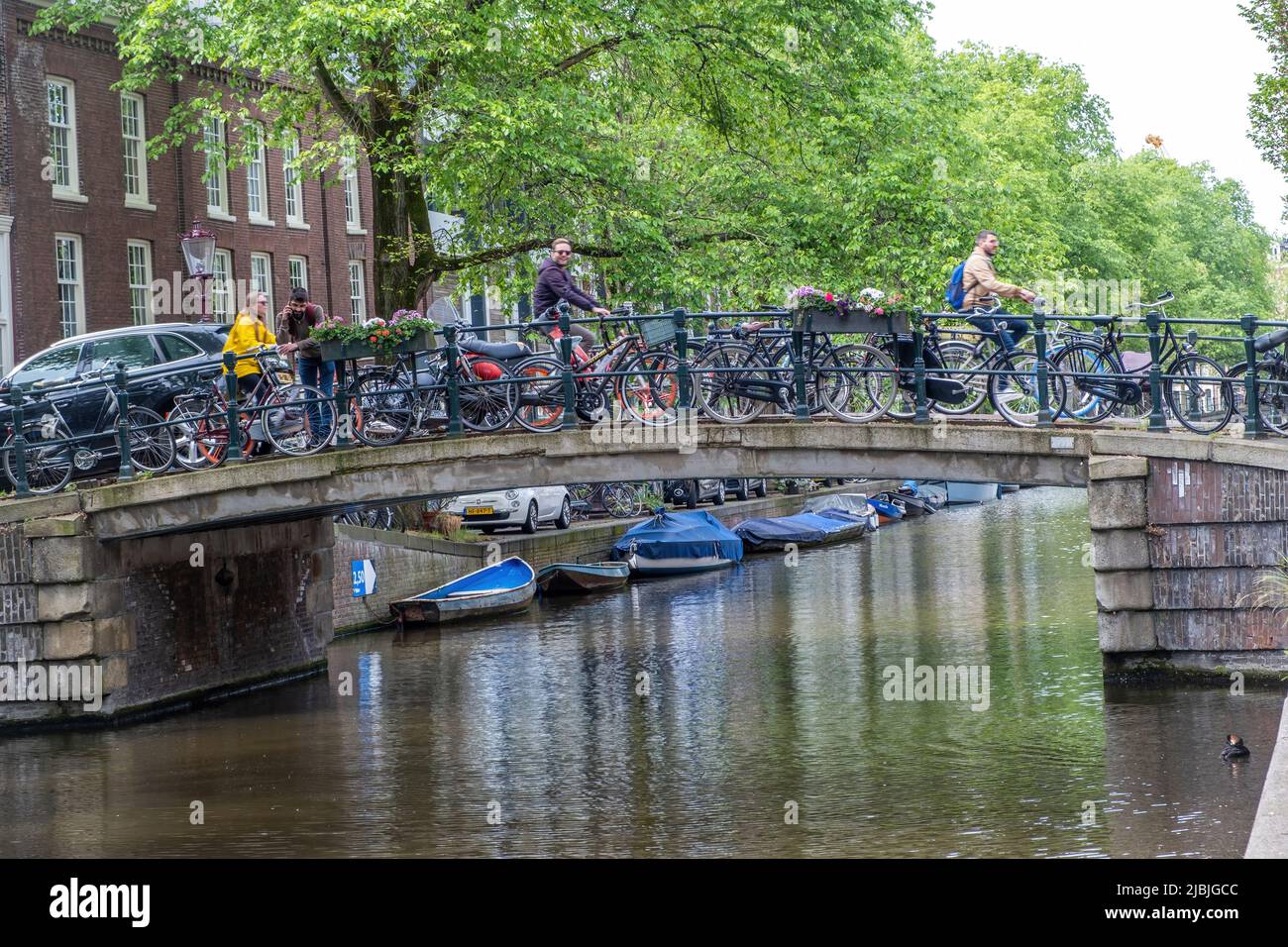 Amsterdam, Niederlande 21. Mai 2022. Die Leute radeln auf einer Brücke, über Kanalwasser, geparkte Fahrräder, festgetäute Boote. Traditionelle Häuser in der holländischen Hauptstadt Stockfoto