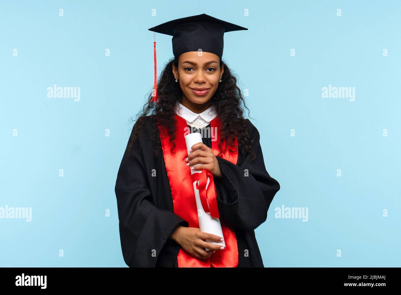 Dunkelhäutige Frau Absolvent trägt Zeremonie Robe und Graduation Cap Holding Zertifikat auf blauem Hintergrund. Mädchen feiert Abschluss und bekommen D Stockfoto