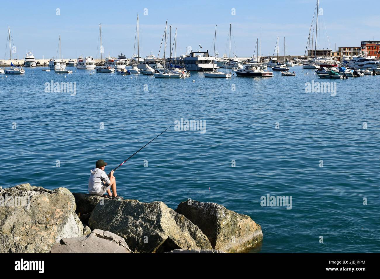Ein Junge (11-12 Jahre alt), der auf den Felsen des alten Seedorfes mit dem Hafen im Hintergrund fischt, Santa Margherita Ligure, Genua, Ligurien, Italien Stockfoto