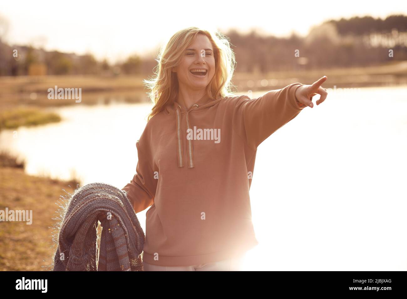 Glückliche, schöne blonde Frau, die an einem kühlen sonnigen Tag allein am Flussufer entlang läuft, nach vorne zeigt und Frieden und Freiheit genießt. Wochenende im Freien Stockfoto