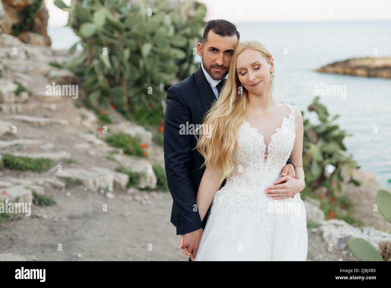 Porträt einer jungen blonden Braut in weißem Kleid und Brunet Bräutigam in Anzug umarmen und halten die Hände auf felsigen Küste in Italien, Seestücke Hintergrund Stockfoto