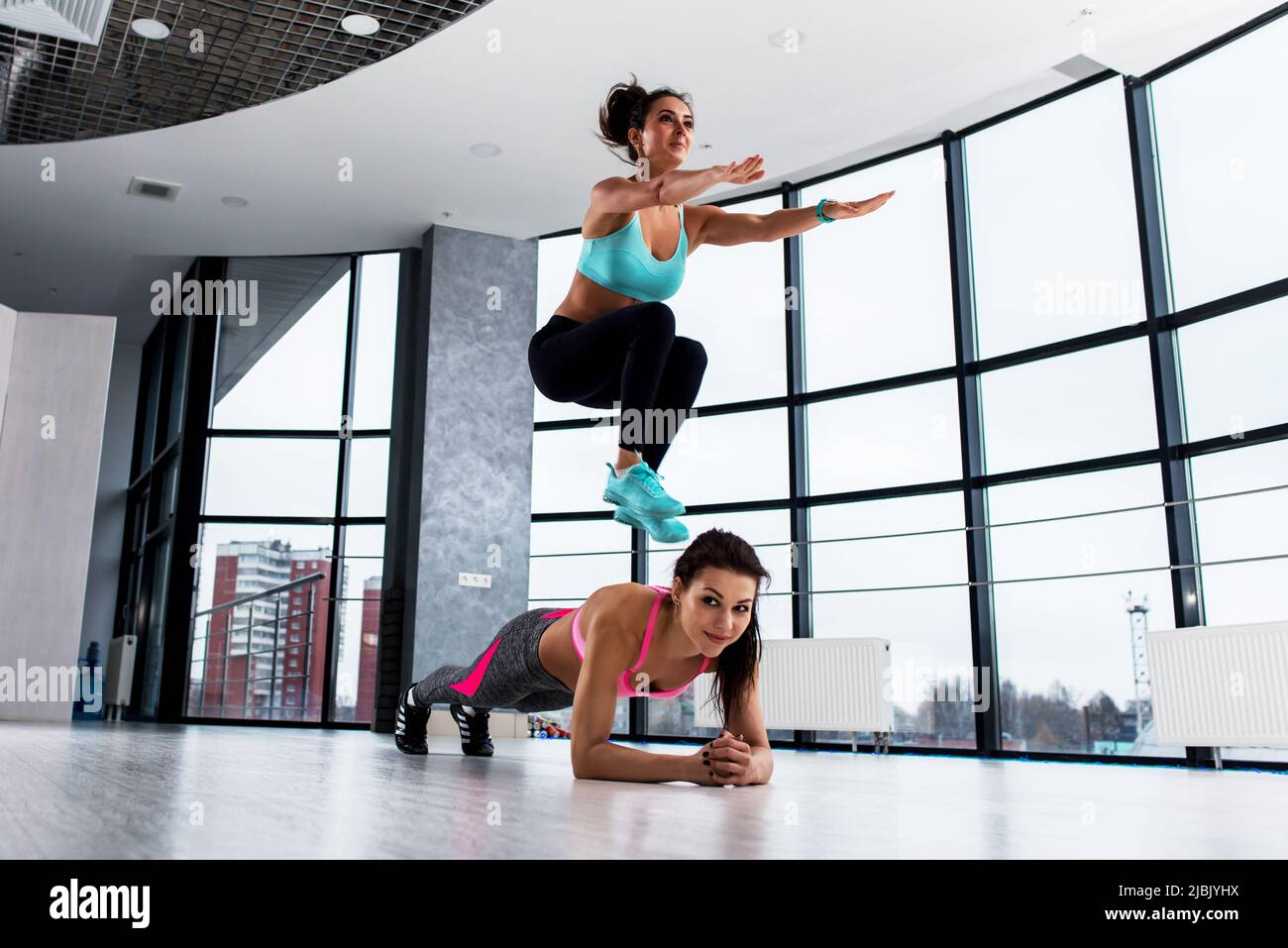 Zwei sportliche kaukasische Mädchen, die ein Partner Circuit Workout absolvieren, die erste Frau hält eine Plankenposition, während die Partnerin im Sport seitlich über sie hockt Stockfoto