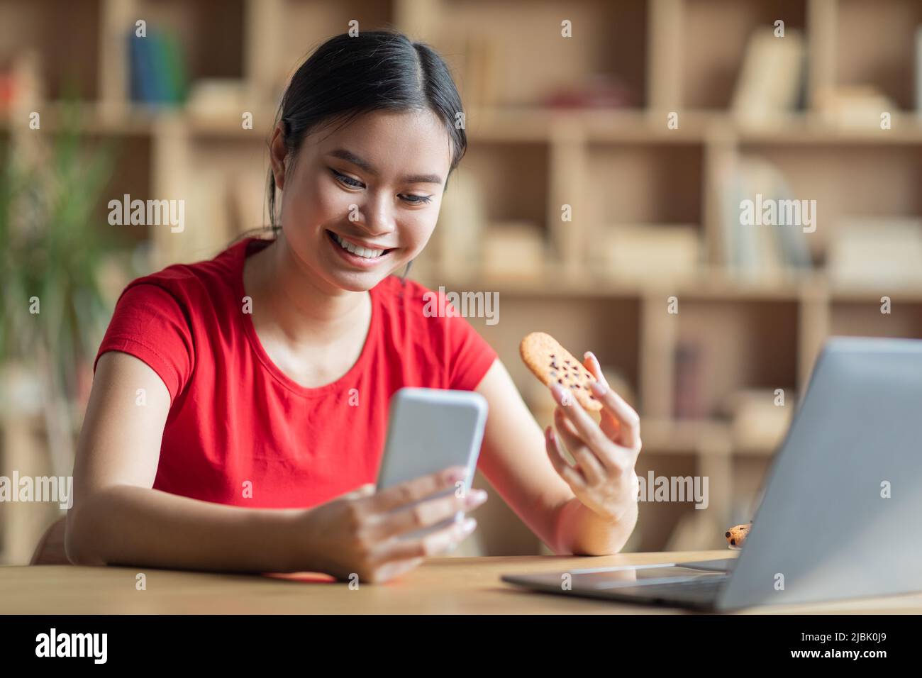 Zufriedene teen koreanische Mädchen Blick auf Telefon, halten Cookie am Tisch mit pc im Zimmer Innenraum Stockfoto
