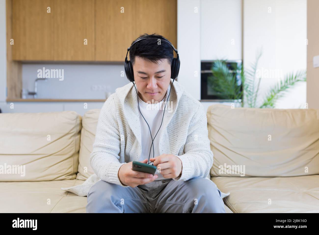 Ein junger, fröhlicher asiatischer Kerl hört Musik in Kopfhörern und sitzt zu Hause auf dem Sofa. Ein Mann allein auf der Couch fühlt sich gut an. Glücklicher Rüde mit dem Stockfoto