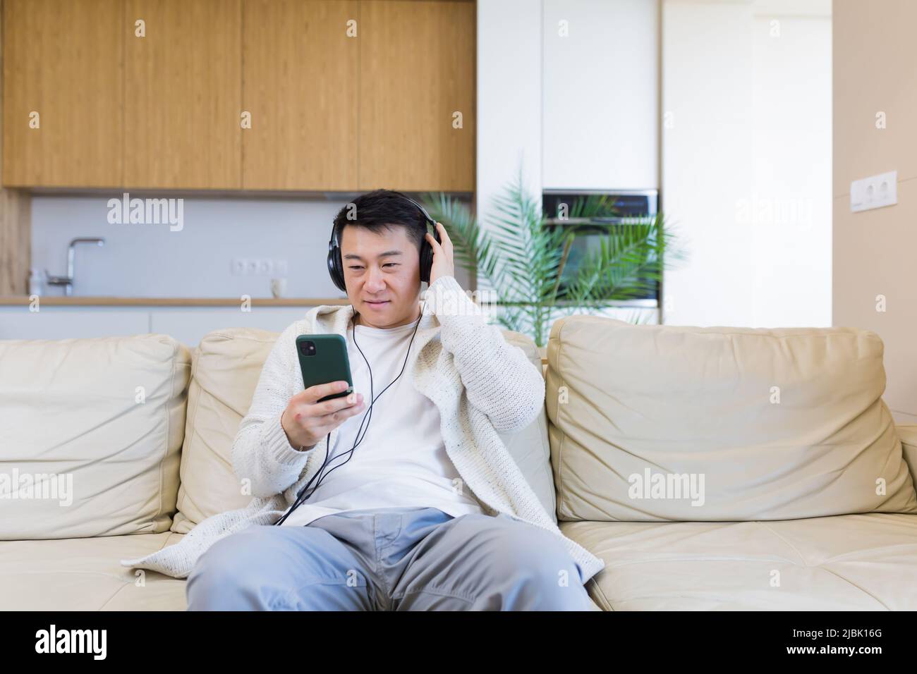 Ein junger, fröhlicher asiatischer Kerl hört Musik in Kopfhörern und sitzt zu Hause auf dem Sofa. Ein Mann allein auf der Couch fühlt sich gut an. Glücklicher Rüde mit dem Stockfoto