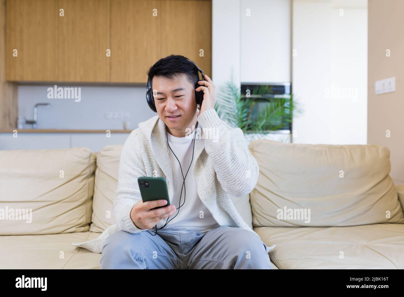 Ein junger, fröhlicher asiatischer Kerl hört Musik in Kopfhörern und sitzt zu Hause auf dem Sofa. Ein Mann allein auf der Couch fühlt sich gut an. Glücklicher Rüde mit dem Stockfoto