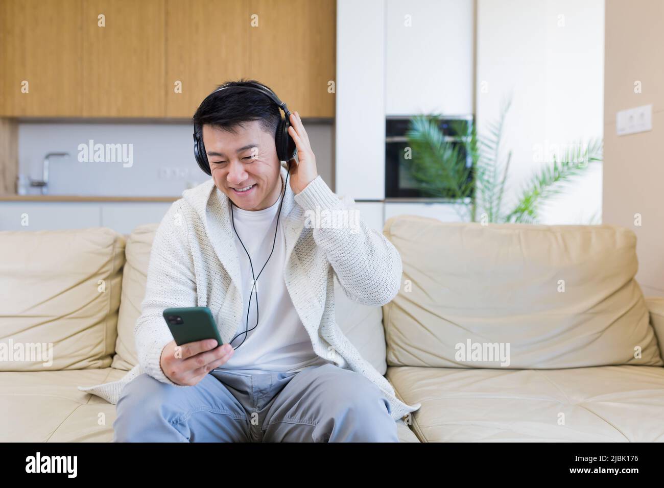 Ein junger, fröhlicher asiatischer Kerl hört Musik in Kopfhörern und sitzt zu Hause auf dem Sofa. Ein Mann allein auf der Couch fühlt sich gut an. Glücklicher Rüde mit dem Stockfoto