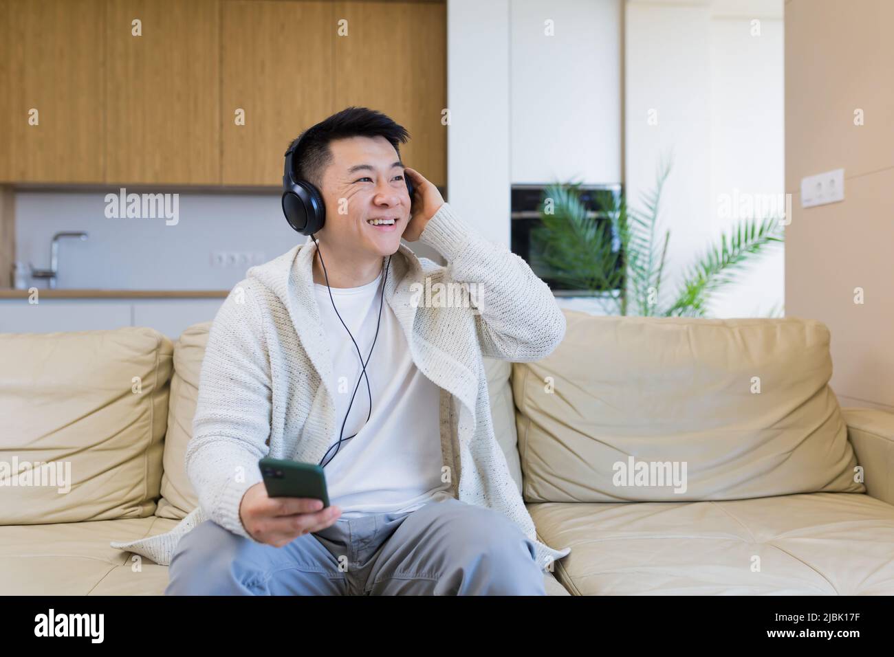 Ein junger, fröhlicher asiatischer Kerl hört Musik in Kopfhörern und sitzt zu Hause auf dem Sofa. Ein Mann allein auf der Couch fühlt sich gut an. Glücklicher Rüde mit dem Stockfoto