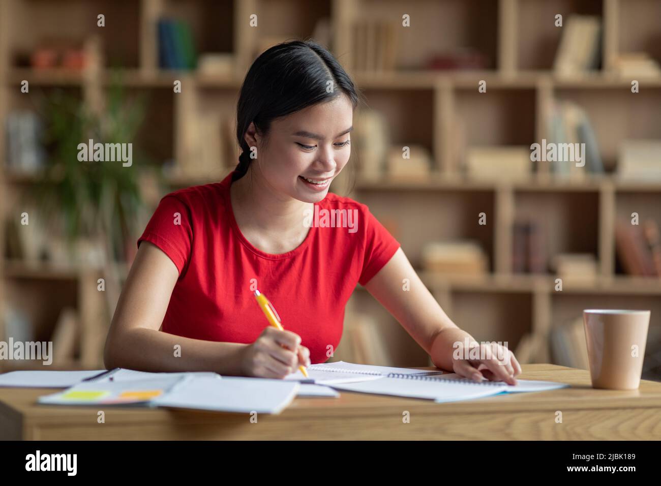 Glückliche hübsche Teenager japanische Frau machen Notizen am Tisch, Studie allein im Wohnzimmer Innenraum, freien Raum Stockfoto