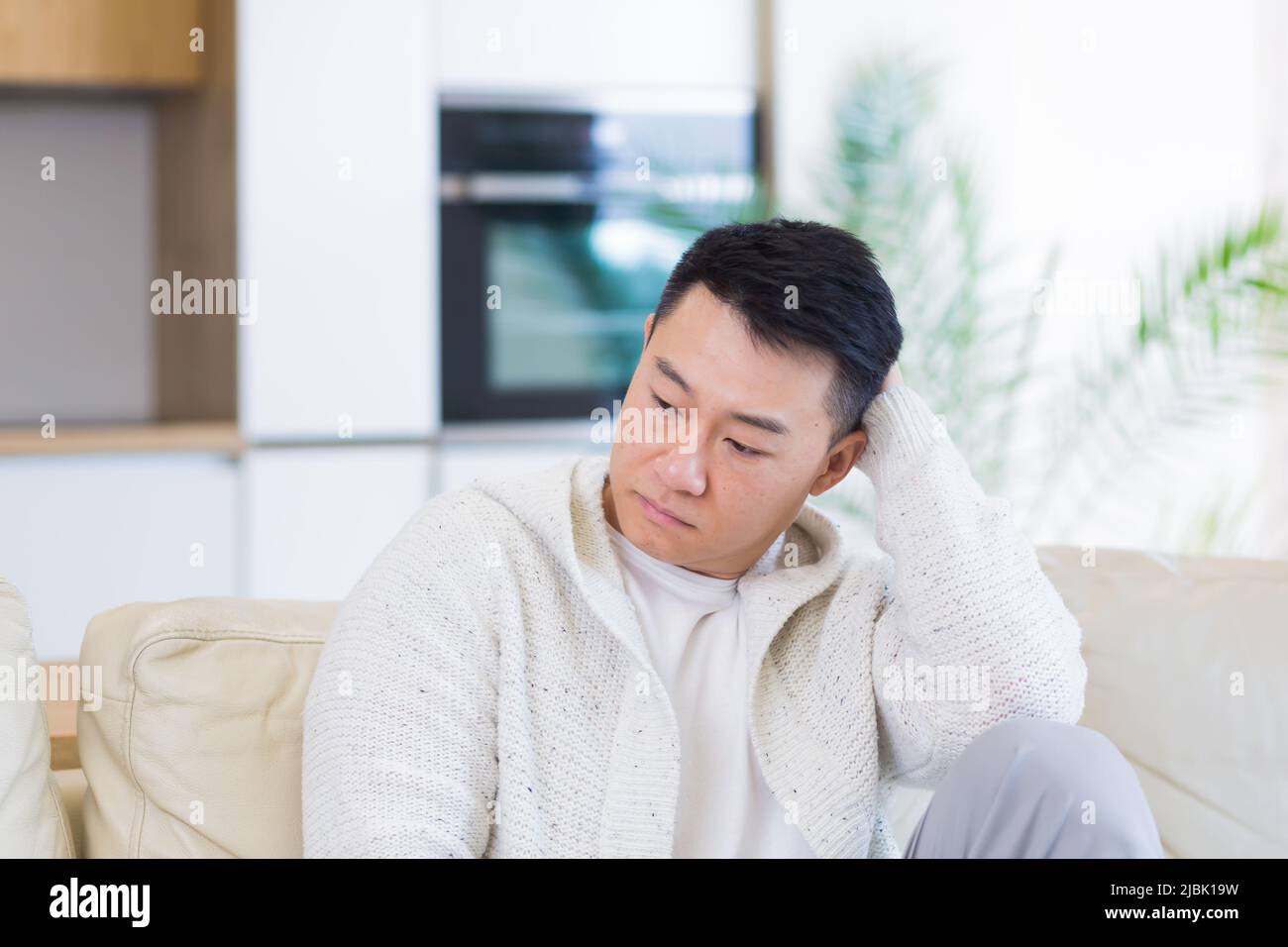 Gelangweilte Freunde junger asiatischer Mann, der allein zu Hause auf dem Sofa sitzt. Vermisst Mann sitzt auf der Couch in der Wohnung. Einsamer Kerl im Wohnzimmer. Ich hatte mir Sorgen gemacht Stockfoto