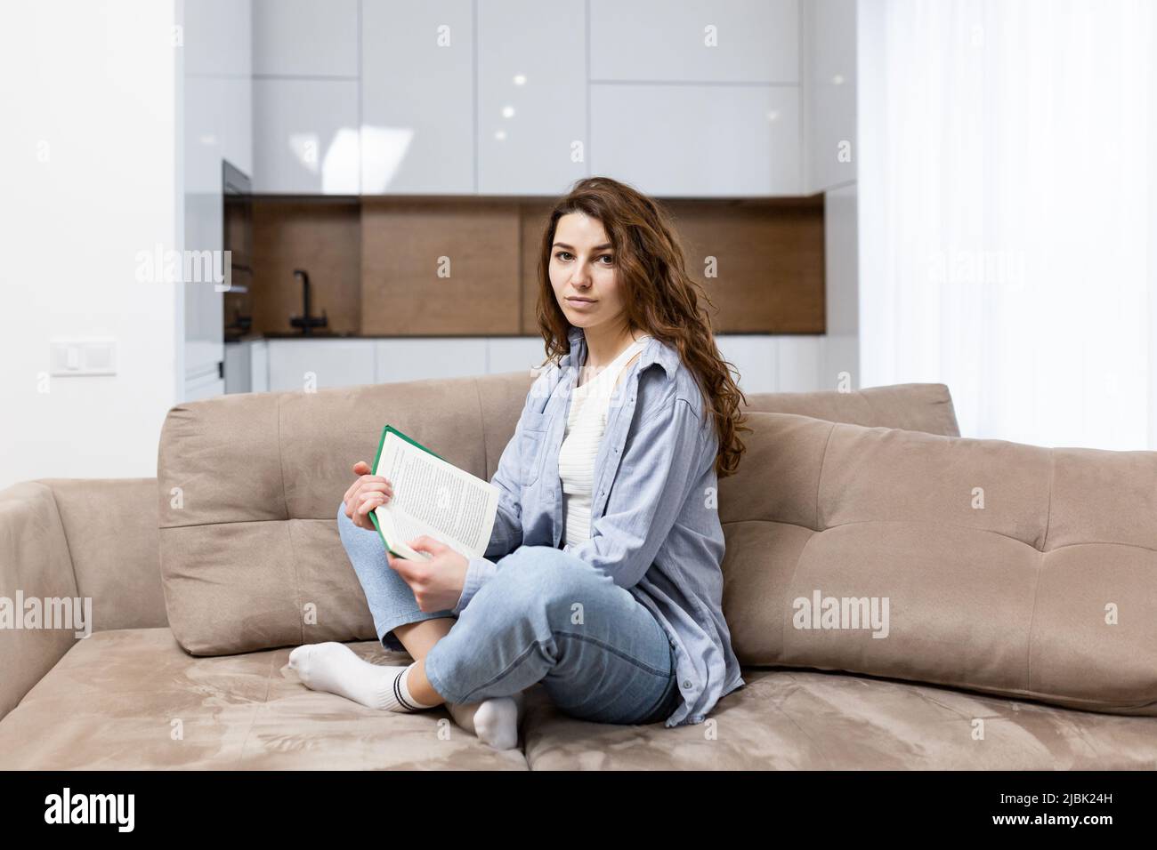 Junge schöne Studentin liest ein Buch, studiert, sitzt zu Hause auf der Couch, bereitet sich auf die Prüfung vor, lächelt, schaut auf die Kamera Stockfoto