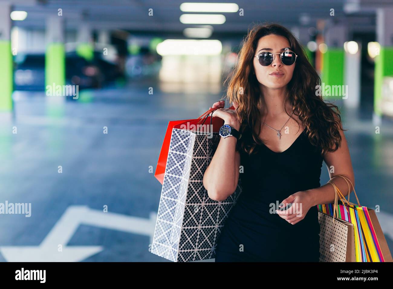 Junge schöne glückliche Frau geht in der Unterführung des Einkaufszentrums zu ihrem Auto mit Einkaufstaschen, Geschenken, Shopaholic Stockfoto