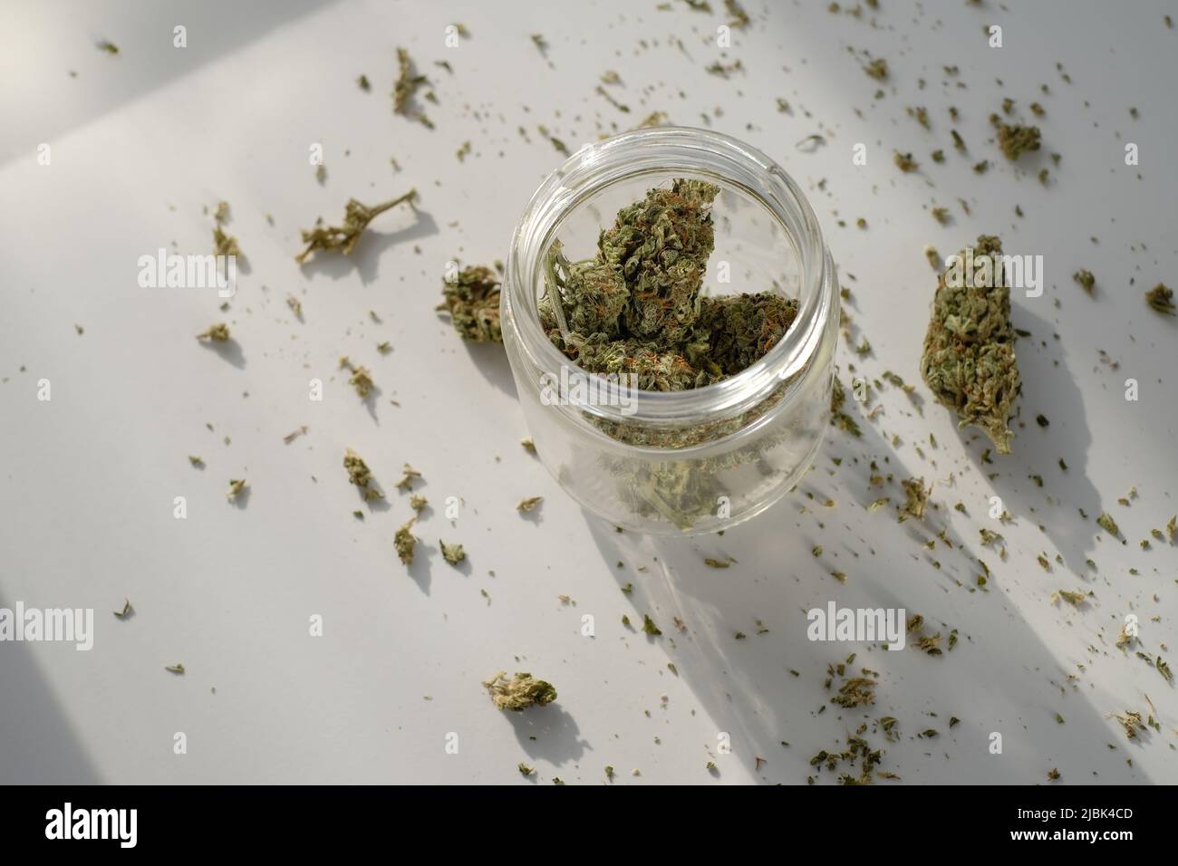 Unkrautladen. Glas mit getrockneten Cannabisknospen auf weißem Hintergrund mit zerdrücktem Marihuana. Stockfoto