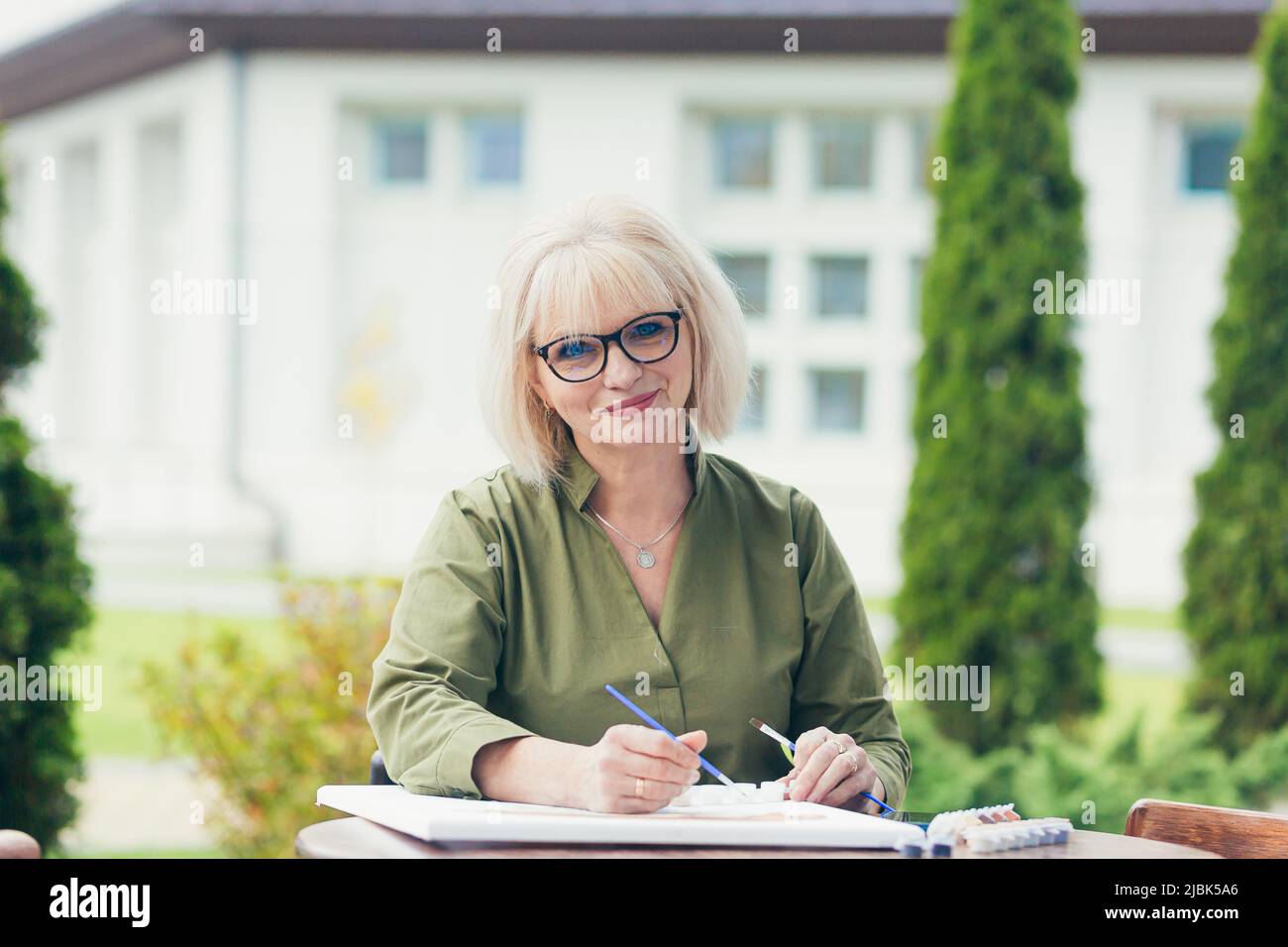 Ältere schöne blonde Frau sitzt an einem Tisch auf der Straße, malt ein Bild, lächelnd, glücklich Stockfoto