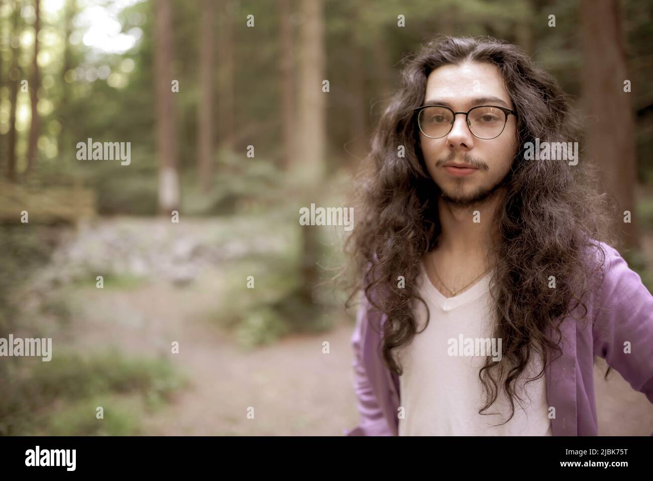 Ein hübscher junger Mann mit langen Haaren steht abseits der Mitte im Wald und blickt direkt auf die Kamera. Unfokussierte Hintergrundkopie. Stockfoto