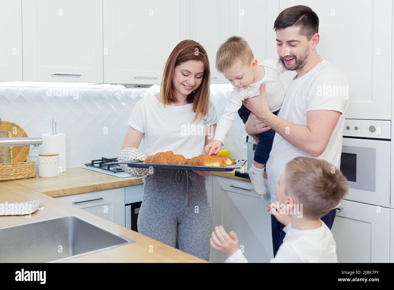 Familienfrühstück, Mama, Papa, zwei Kinder, Jungen, die von Mama gebackene Croissants aßen, Kaffee und frischen Saft tranken, lächelten und sich zu Hause in der Kitch umarmten Stockfoto