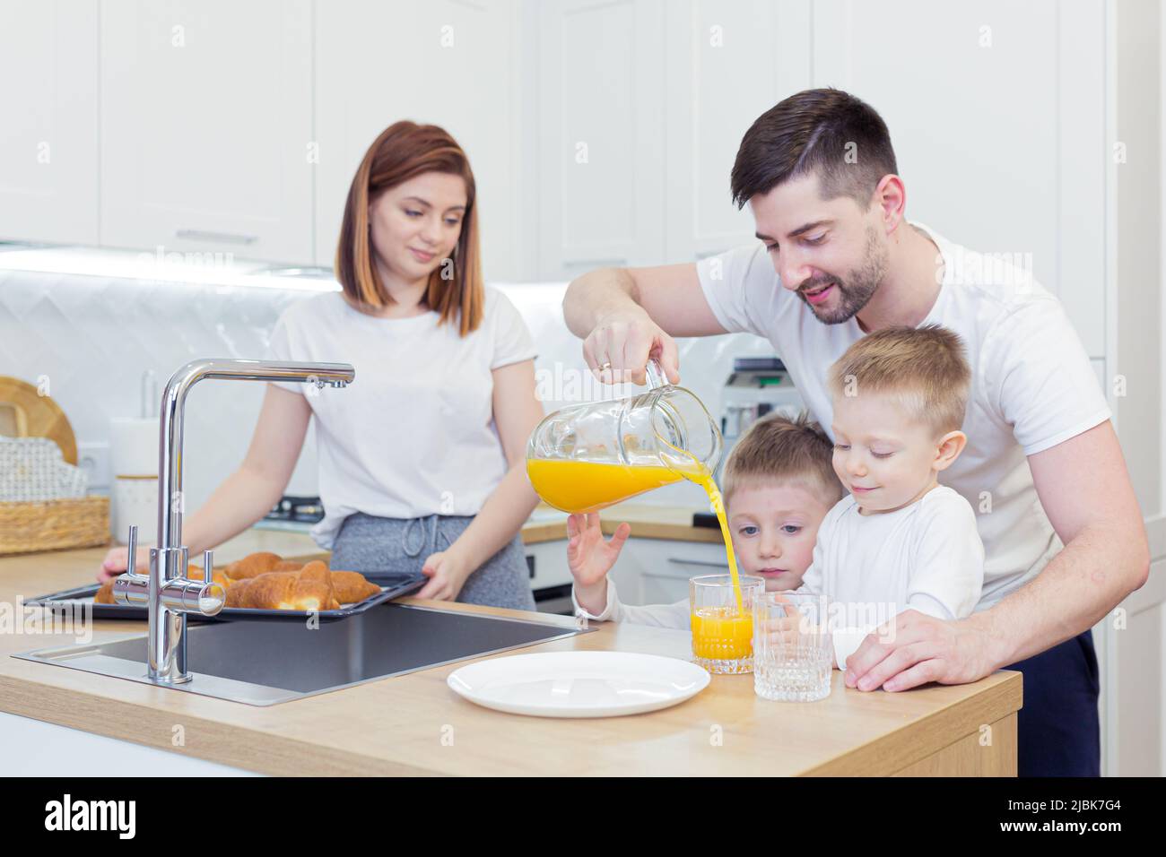 Familienfrühstück, Mama, Papa, zwei Kinder, Jungen, die von Mama gebackene Croissants aßen, Kaffee und frischen Saft tranken, lächelten und sich zu Hause in der Kitch umarmten Stockfoto