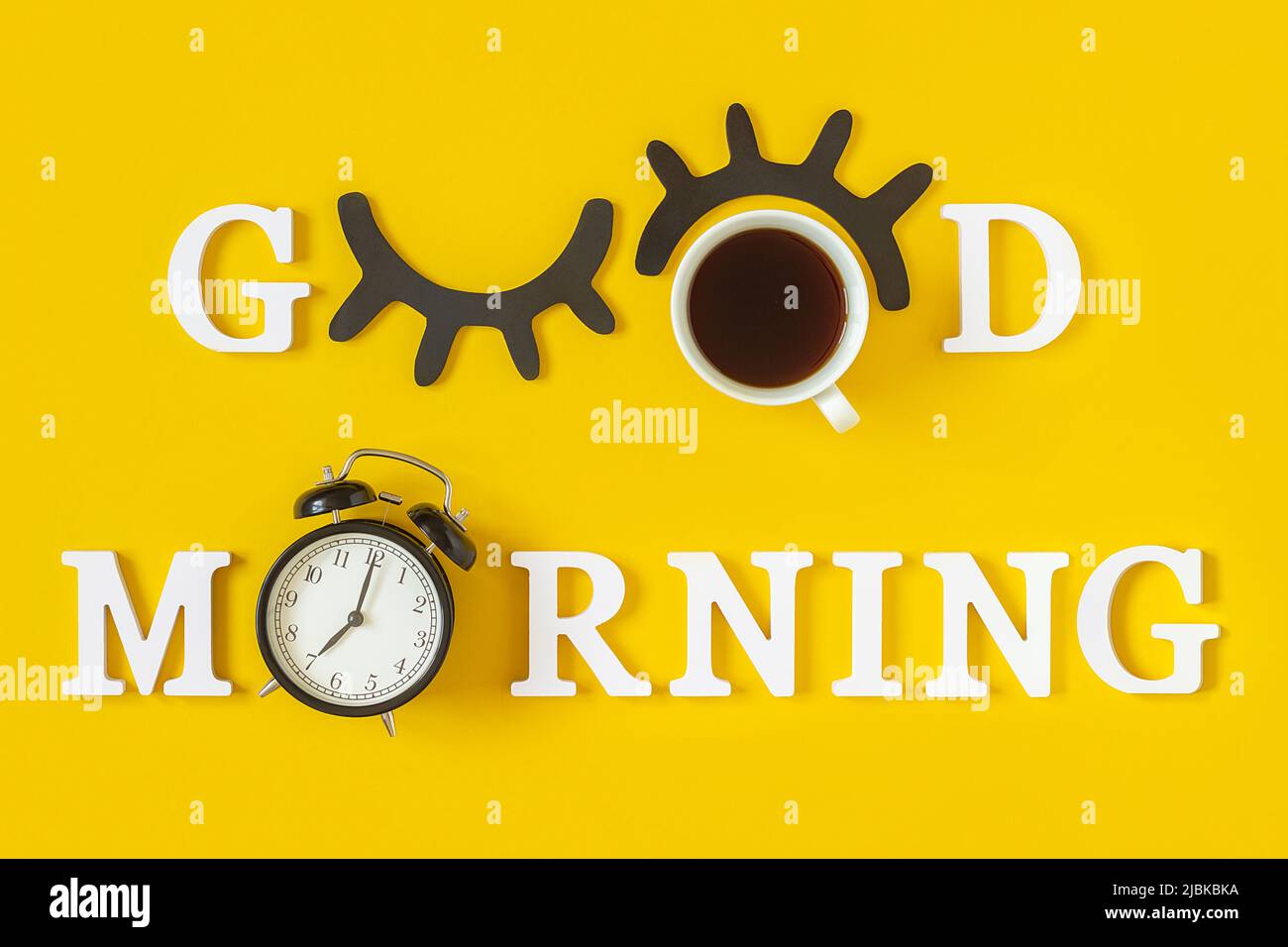 Kreatives Good Morning-Konzept. Weiße Buchstaben, schwarze Wimpern, Wecker und eine Tasse Kaffee auf gelbem Hintergrund. Draufsicht, Flat Lay. Grußkarte. Stockfoto