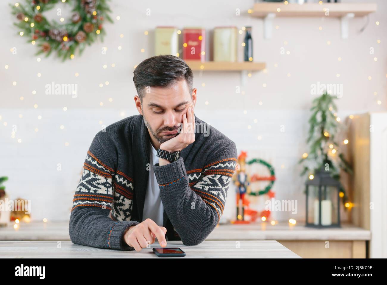 Ein Mann mit einem Bart ist gelangweilt und ist zu Weihnachten allein zu Hause und schaut in Depressionen auf das Telefon Stockfoto